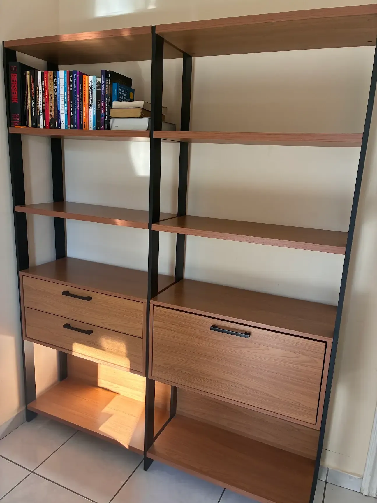 Estante modulada para livros com estrutura preta, prateleiras amadeiradas e gaveteiro